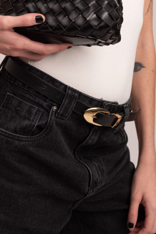 Plano médio de modelo segurando bolsa preta, vestindo jeans escuro e regata branca. O foco central da fotografia é o cinto de couro preto, evidenciando o brilho da fivela dourada e o acabamento premium do couro legítimo contra o fundo branco.