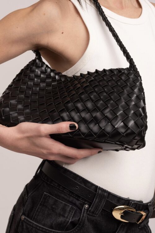 Mulher segura a bolsa de couro preta Kaya entre o busto e a cintura. Detalhe para a textura trançada da alça e fechamento em zíper de metal. Fundo neutro com foco no acessório.