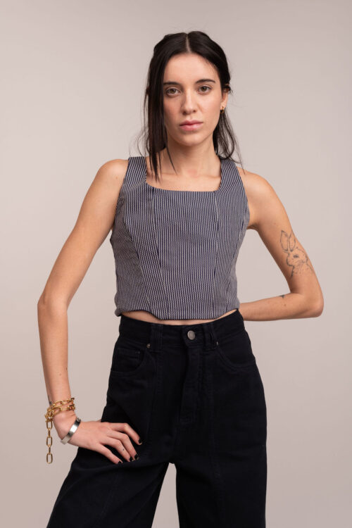 Modelo de cabelos escuros veste o corset de alfaiataria estruturado Jade combinado com calça preta. A foto, em plano americano (até os joelhos) sobre fundo neutro, destaca a modelagem da peça em uma pose fashion e moderna.