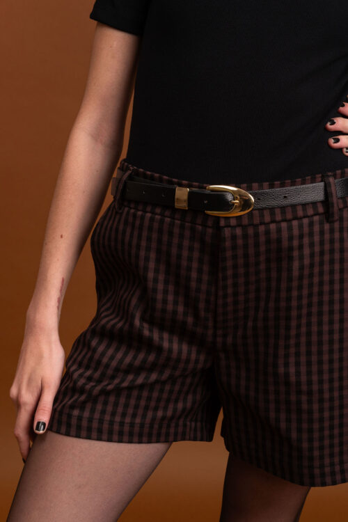 Modelo veste blusa preta e shorts xadrez, utilizando o cinto de couro preto com fivela dourada para marcar a silhueta. Retrato enquadrado do tronco às coxas em cenário com fundo marrom.